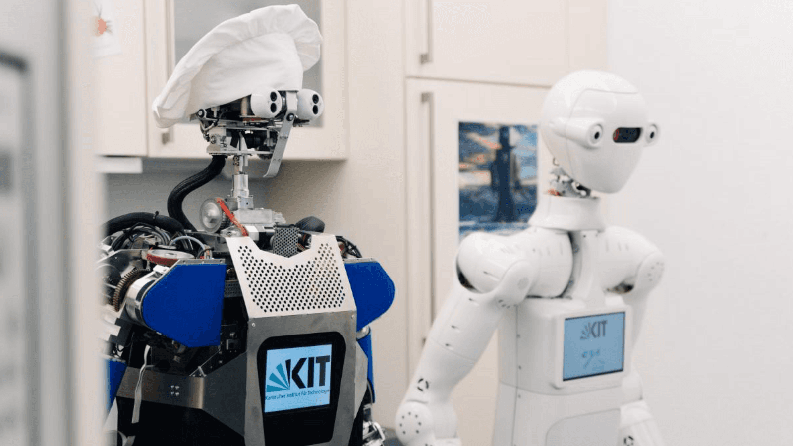 Two humanoid research robots in a kitchen, one wearing a chef's hat, showing AI moving into physical work